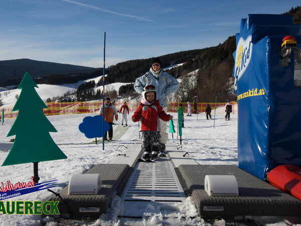 Ein Kind mit Helm steht in Begleitung eines Erwachsenen an einem Förderband-Skilift in einem verschneiten Skigebiet mit Sicherheitsbarrieren und dekorativen Aussparungen.