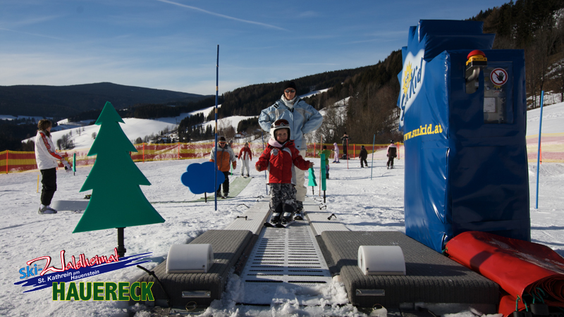 Ein Kind mit Helm steht in Begleitung eines Erwachsenen an einem Förderband-Skilift in einem verschneiten Skigebiet mit Sicherheitsbarrieren und dekorativen Aussparungen.