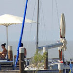 Menschen entspannen sich unter Sonnenschirmen auf einem Steg in der Nähe mehrerer Segelboote, mit ruhigem Wasser und fernen Segelbooten im Hintergrund.