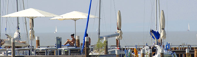 Menschen entspannen sich unter Sonnenschirmen auf einem Steg in der Nähe mehrerer Segelboote, mit ruhigem Wasser und fernen Segelbooten im Hintergrund.