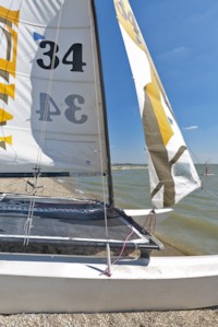 Nahaufnahme eines kleinen Segelboots mit der Nummer 34 auf dem Segel, das an einem Kiesstrand neben ruhigem Wasser unter einem klaren blauen Himmel liegt.