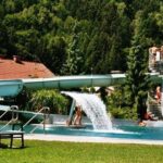 Menschen stehen auf einer Wendeltreppe und warten auf eine Wasserrutsche in einem von Bäumen und Häusern umgebenen Freibad, wobei eine Person auf einer Bank im Vordergrund sitzt.