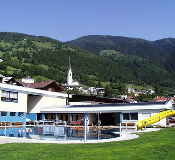 Freibad mit gelber Rutsche neben modernen Gebäuden, eingebettet in eine grüne Hügellandschaft mit einer Kirche und Häusern im Hintergrund.