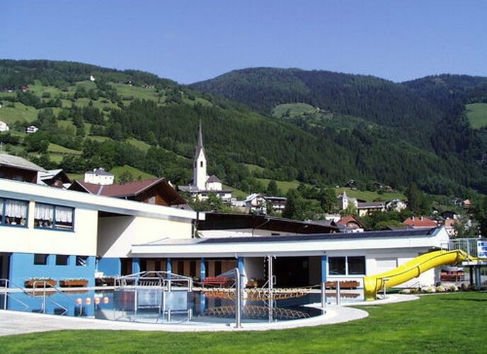Freibad mit gelber Rutsche neben modernen Gebäuden, eingebettet in eine grüne Hügellandschaft mit einer Kirche und Häusern im Hintergrund.