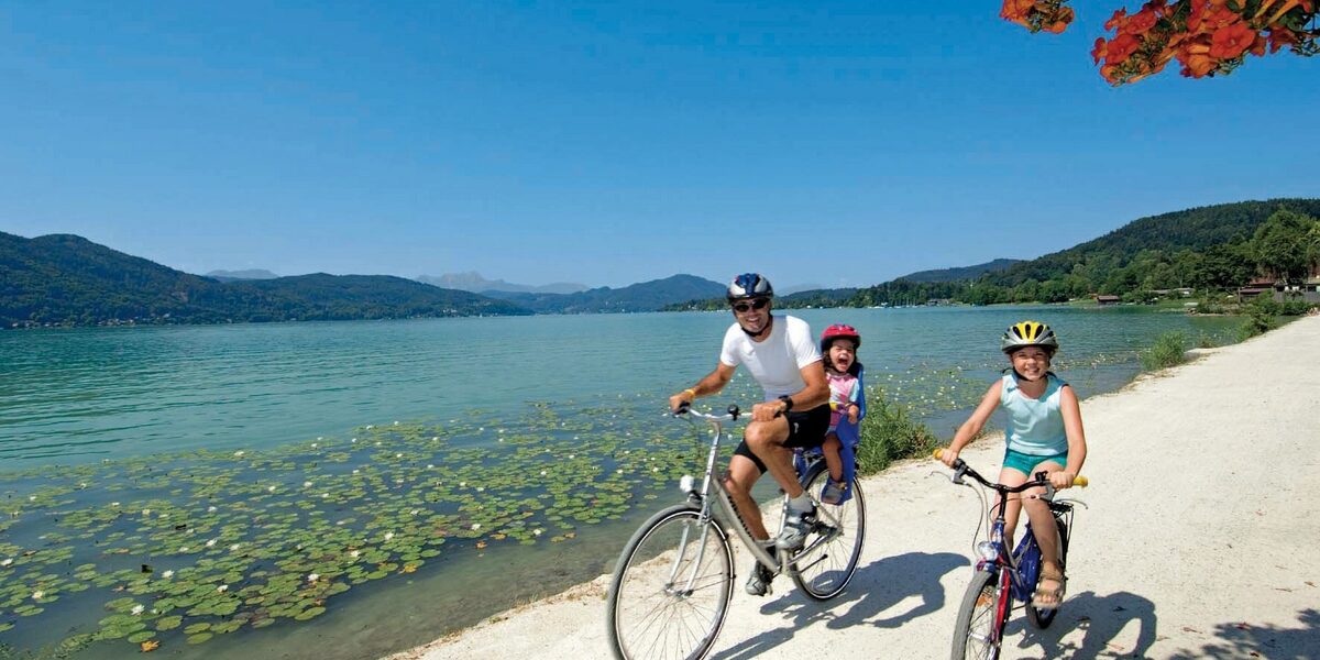 Zwei Erwachsene und ein Kind fahren mit dem Fahrrad auf einem Seeuferweg unter blauem Himmel, mit Bergen und Seerosen im Hintergrund.