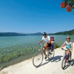 Zwei Erwachsene und ein Kind fahren mit dem Fahrrad auf einem Seeuferweg unter blauem Himmel, mit Bergen und Seerosen im Hintergrund.