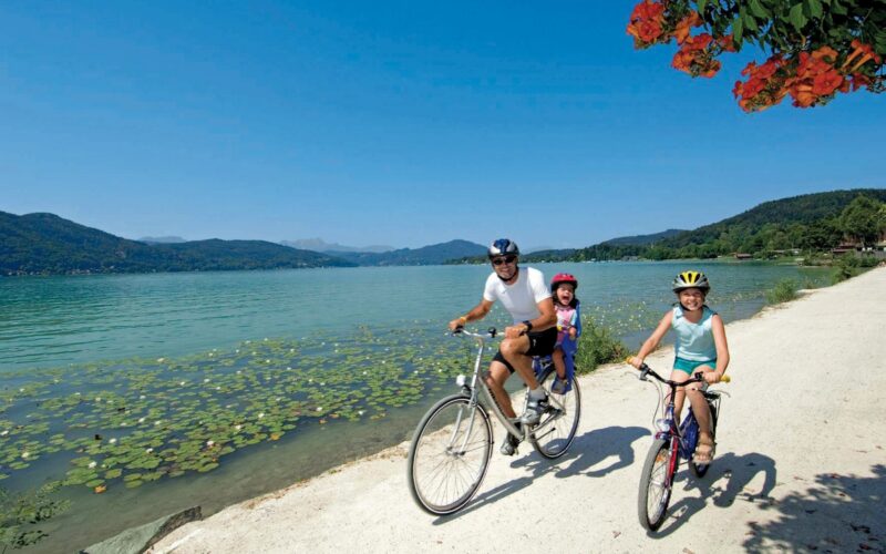 Zwei Erwachsene und ein Kind fahren mit dem Fahrrad auf einem Seeuferweg unter blauem Himmel, mit Bergen und Seerosen im Hintergrund.