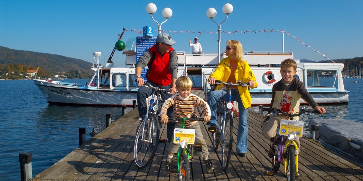 Vier Personen, zwei Erwachsene und zwei Kinder, stehen an einem sonnigen Tag mit Fahrrädern auf einem hölzernen Steg neben einem festgemachten Boot, mit Hügeln und Wasser im Hintergrund.