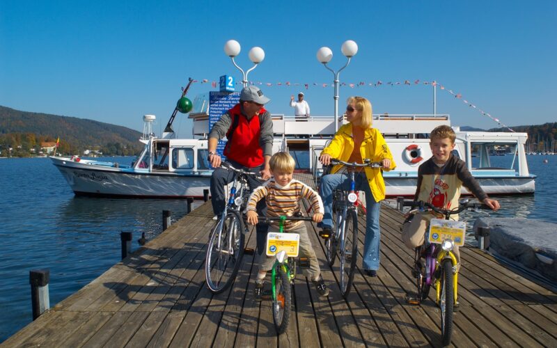 Vier Personen, zwei Erwachsene und zwei Kinder, stehen an einem sonnigen Tag mit Fahrrädern auf einem hölzernen Steg neben einem festgemachten Boot, mit Hügeln und Wasser im Hintergrund.