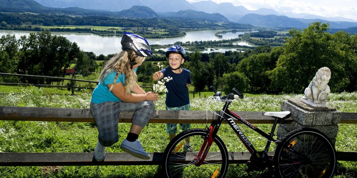 Zwei Kinder mit Helmen sitzen auf einem Holzzaun neben ihren Fahrrädern und teilen sich einen Snack mit einem malerischen See und einer Berglandschaft im Hintergrund.