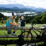 Zwei Kinder mit Helmen sitzen auf einem Holzzaun neben ihren Fahrrädern und teilen sich einen Snack mit einem malerischen See und einer Berglandschaft im Hintergrund.