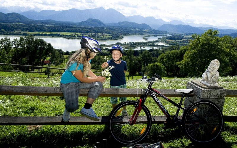 Zwei Kinder mit Helmen sitzen auf einem Holzzaun neben ihren Fahrrädern und teilen sich einen Snack mit einem malerischen See und einer Berglandschaft im Hintergrund.