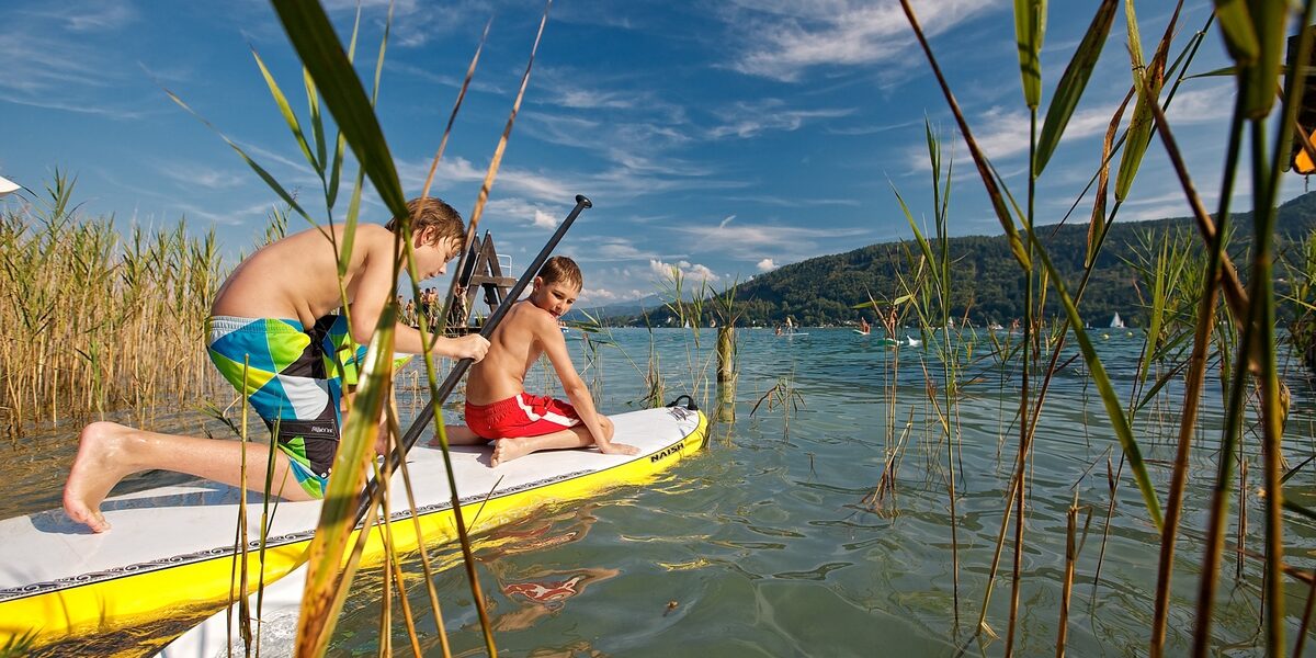 Zwei Jungen in Badehosen stehen auf einem gelben Paddelbrett im seichten Wasser in der Nähe von Schilf, mit Bergen und blauem Himmel im Hintergrund.