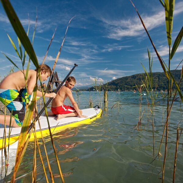 Zwei Jungen in Badehosen stehen auf einem gelben Paddelbrett im seichten Wasser in der Nähe von Schilf, mit Bergen und blauem Himmel im Hintergrund.