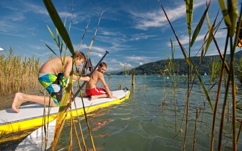Zwei Jungen in Badehosen stehen auf einem gelben Paddelbrett im seichten Wasser in der Nähe von Schilf, mit Bergen und blauem Himmel im Hintergrund.