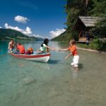 Vier Personen an einem Seeufer: zwei in einem Ruderboot, einer, der das Boot zieht, und einer am Ufer, der zu Hilfe eilt, mit einer Hütte und Bäumen im Hintergrund.