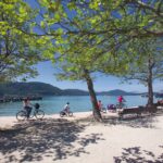 An einem sonnigen Tag fahren Menschen mit ihren Fahrrädern einen von Bäumen gesäumten Weg an einem See entlang, mit Bergen und einem Pier im Hintergrund.