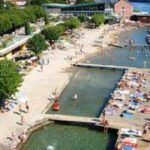 Ein überfüllter Badeplatz am See mit Sonnenanbetern auf Betonplattformen, schwimmenden Menschen und Bäumen am Ufer.