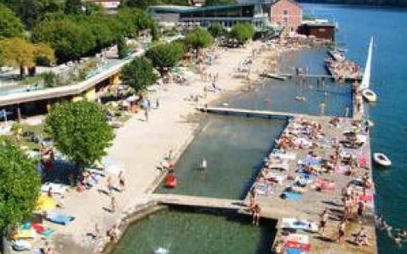 Ein überfüllter Badeplatz am See mit Sonnenanbetern auf Betonplattformen, schwimmenden Menschen und Bäumen am Ufer.