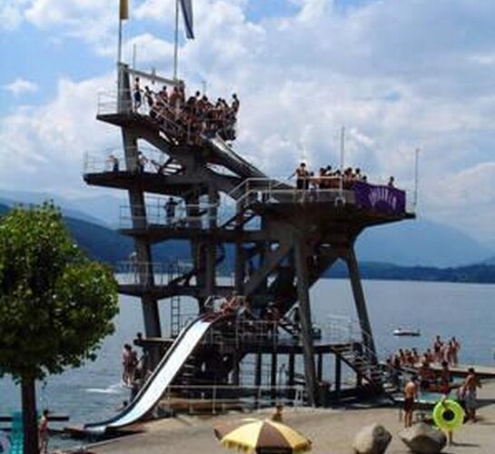 Ein hoher, mehrstöckiger Sprungturm, auf dem auf jeder Ebene Menschen stehen, überblickt einen See mit Schwimmern, einer Wasserrutsche und einem Sandstrand im Vordergrund.