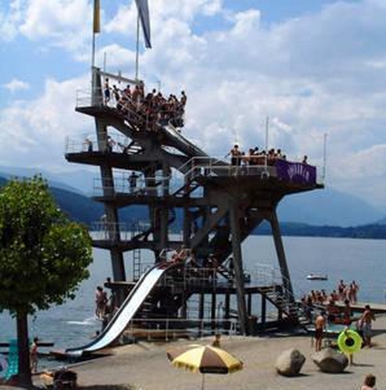 Ein hoher, mehrstöckiger Sprungturm, auf dem auf jeder Ebene Menschen stehen, überblickt einen See mit Schwimmern, einer Wasserrutsche und einem Sandstrand im Vordergrund.