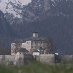 Eine große Steinburg mit vielen Fenstern steht auf einem befestigten Sockel, mit schneebedeckten Bergen im Hintergrund.