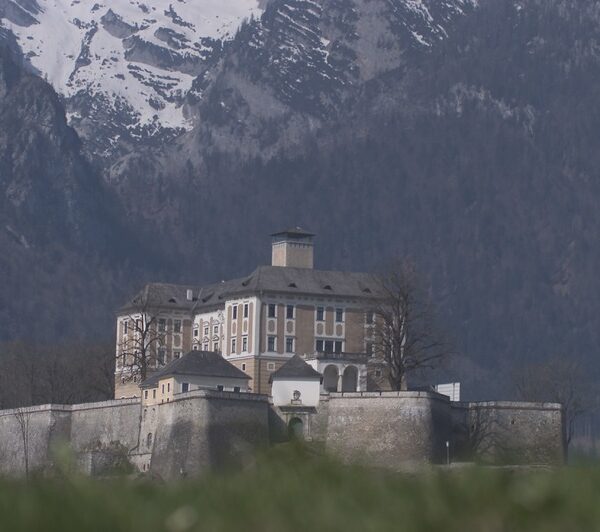 Eine große Steinburg mit vielen Fenstern steht auf einem befestigten Sockel, mit schneebedeckten Bergen im Hintergrund.