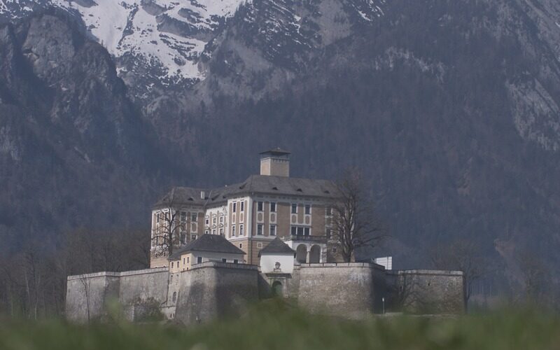 Eine große Steinburg mit vielen Fenstern steht auf einem befestigten Sockel, mit schneebedeckten Bergen im Hintergrund.