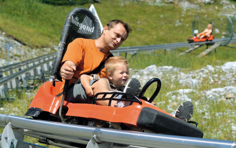 Ein Erwachsener und ein Kind fahren an einem sonnigen Tag gemeinsam auf einer Alpenachterbahn, während im Hintergrund ein weiterer Fahrer zu sehen ist.