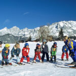 Eine Gruppe von Kindern auf Skiern steht in einer Reihe vor einem Skilehrer auf einer verschneiten Piste, mit Bergen im Hintergrund unter einem klaren blauen Himmel.