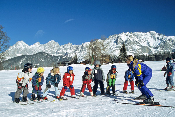Eine Gruppe von Kindern auf Skiern steht in einer Reihe vor einem Skilehrer auf einer verschneiten Piste, mit Bergen im Hintergrund unter einem klaren blauen Himmel.