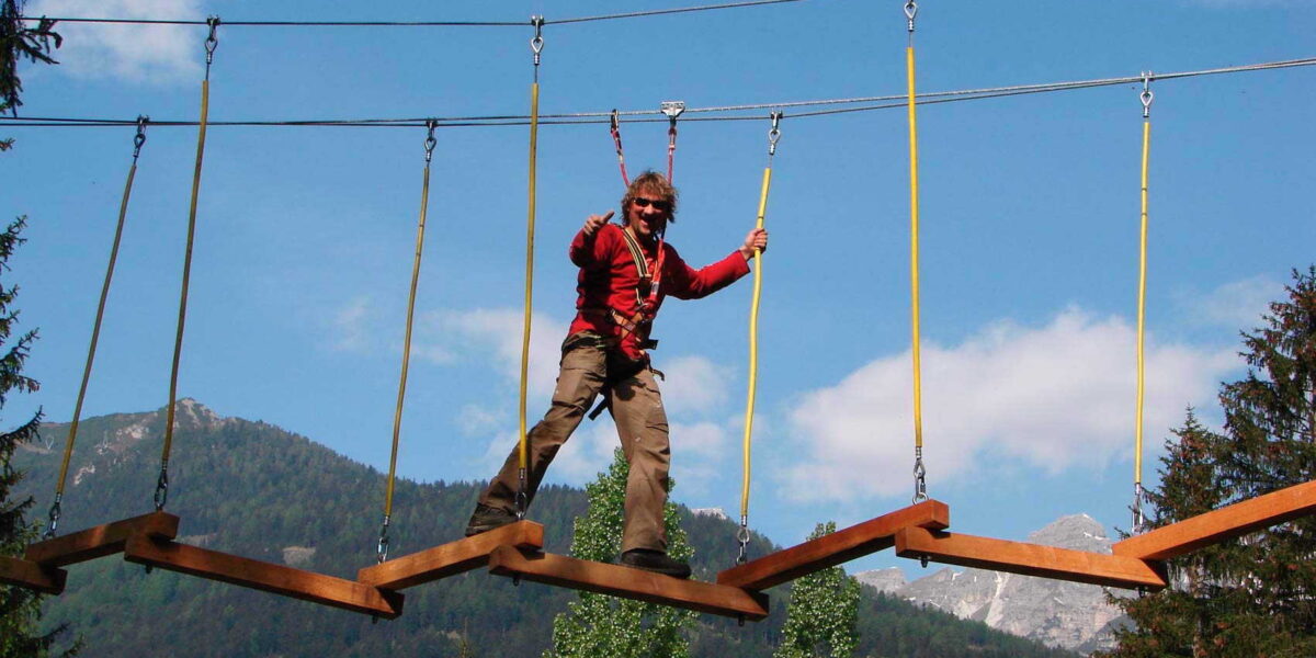 Eine Person mit einem Klettergurt überquert eine hängende Zickzack-Holzbrücke in einem Outdoor-Abenteuerpark mit Bergen und Bäumen im Hintergrund.