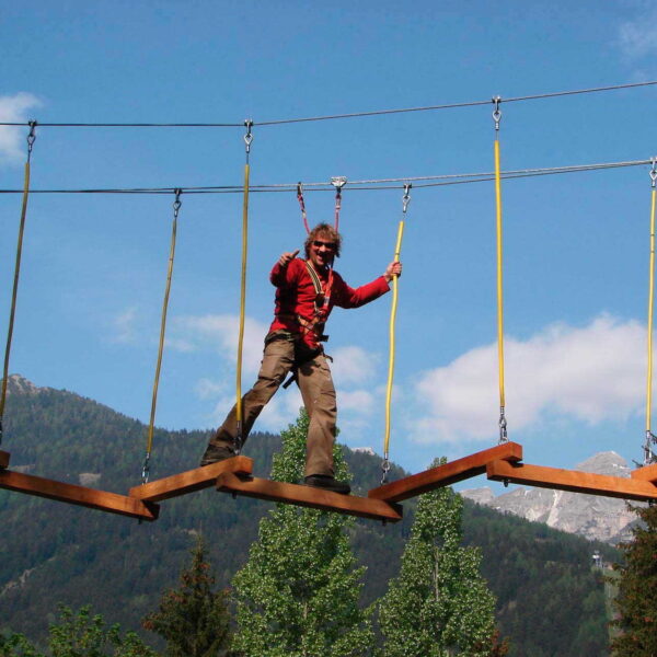 Eine Person mit einem Klettergurt überquert eine hängende Zickzack-Holzbrücke in einem Outdoor-Abenteuerpark mit Bergen und Bäumen im Hintergrund.