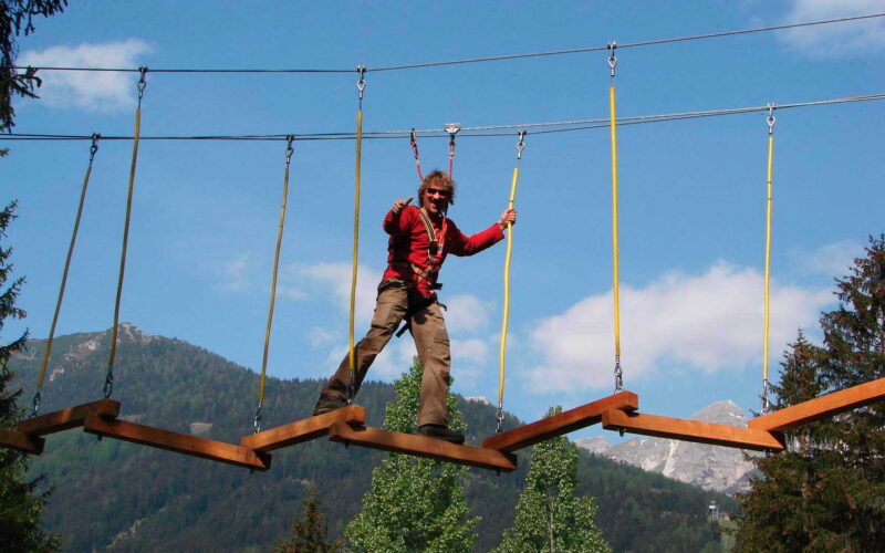 Eine Person mit einem Klettergurt überquert eine hängende Zickzack-Holzbrücke in einem Outdoor-Abenteuerpark mit Bergen und Bäumen im Hintergrund.