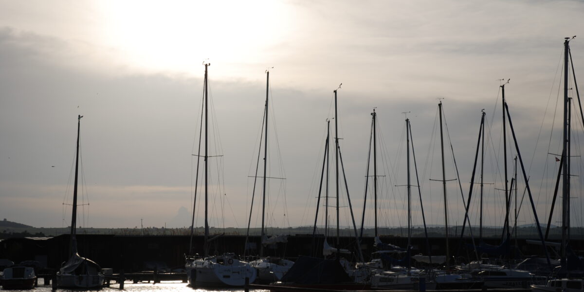 In einem Jachthafen liegen Segelboote vor Anker, deren hohe Masten sich gegen den wolkenverhangenen Himmel abzeichnen, während die Sonne tief am Horizont steht.