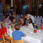 Eine Gruppe von Kindern sitzt bei einer Party um einen dekorierten Tisch herum, Erwachsene stehen in der Nähe; auf dem Tisch stehen Partygeschenke und Getränke.