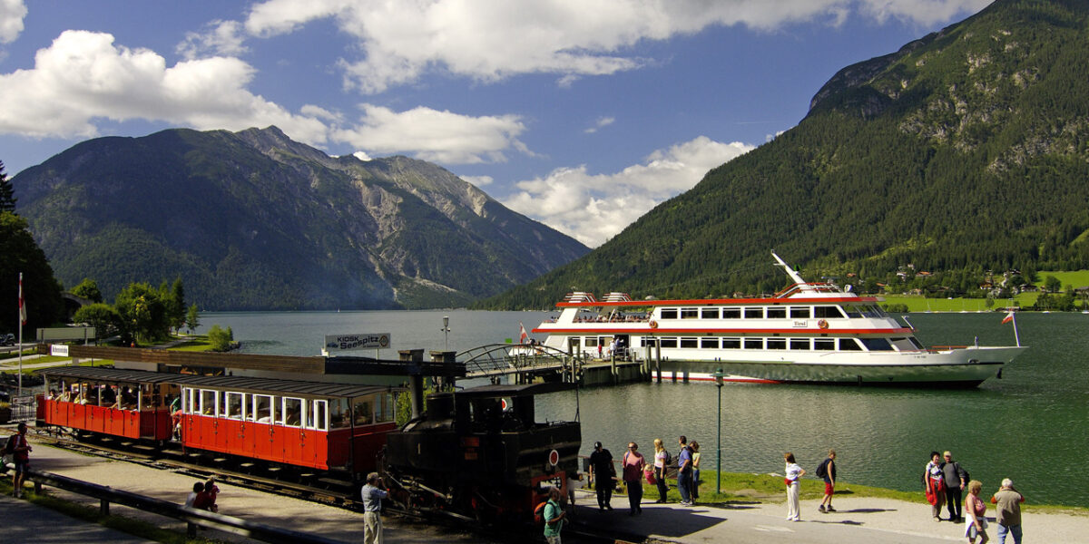 Ein historischer Zug parkt an einem Bahnhof am Seeufer, in dessen Nähe sich Menschen aufhalten; ein großes weiß-rotes Boot liegt auf dem grünen See vor Anker, umgeben von Bergen unter einem teilweise bewölkten Himmel.