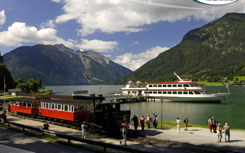 Ein historischer Zug parkt an einem Bahnhof am Seeufer, in dessen Nähe sich Menschen aufhalten; ein großes weiß-rotes Boot liegt auf dem grünen See vor Anker, umgeben von Bergen unter einem teilweise bewölkten Himmel.
