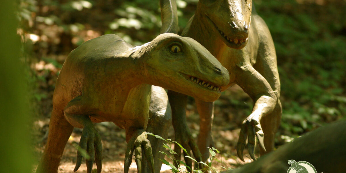 Zwei realistische Dinosauriermodelle sind in einem Wald zu sehen, in dem das Sonnenlicht durch die Bäume fällt. Das "Styrassic Park"-Logo ist in der unteren rechten Ecke zu sehen.