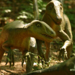 Zwei realistische Dinosauriermodelle sind in einem Wald zu sehen, in dem das Sonnenlicht durch die Bäume fällt. Das "Styrassic Park"-Logo ist in der unteren rechten Ecke zu sehen.