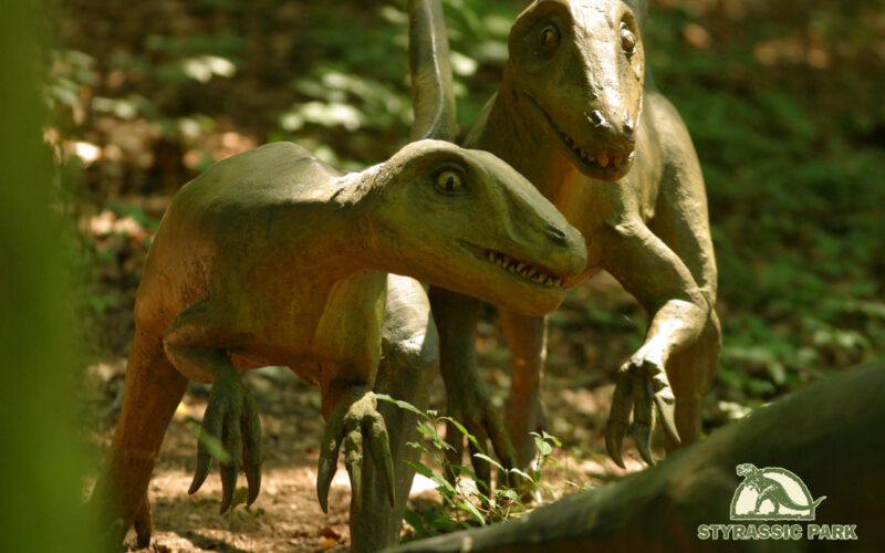 Zwei realistische Dinosauriermodelle sind in einem Wald zu sehen, in dem das Sonnenlicht durch die Bäume fällt. Das "Styrassic Park"-Logo ist in der unteren rechten Ecke zu sehen.