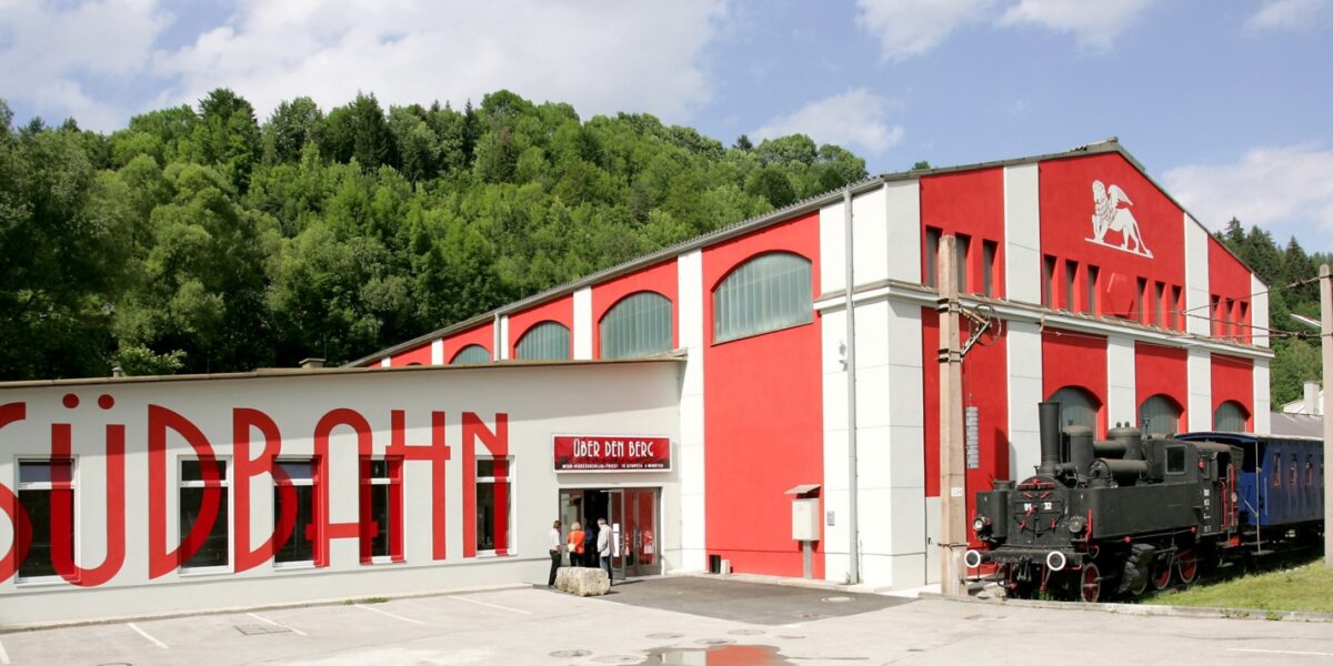 Ein rot-weißes Gebäude mit der Aufschrift "Südbahn", vor dem eine alte schwarze Dampflokomotive und Waggons ausgestellt sind, umgeben von Grünpflanzen.