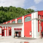 Ein rot-weißes Gebäude mit der Aufschrift "Südbahn", vor dem eine alte schwarze Dampflokomotive und Waggons ausgestellt sind, umgeben von Grünpflanzen.