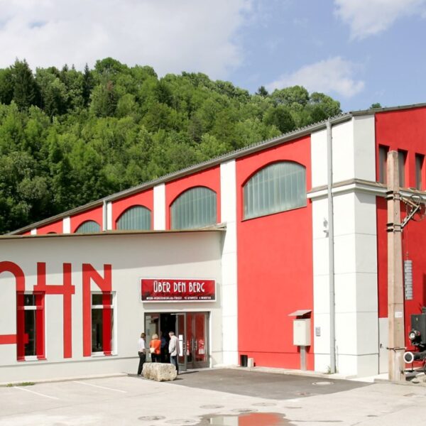 Ein rot-weißes Gebäude mit der Aufschrift "Südbahn", vor dem eine alte schwarze Dampflokomotive und Waggons ausgestellt sind, umgeben von Grünpflanzen.