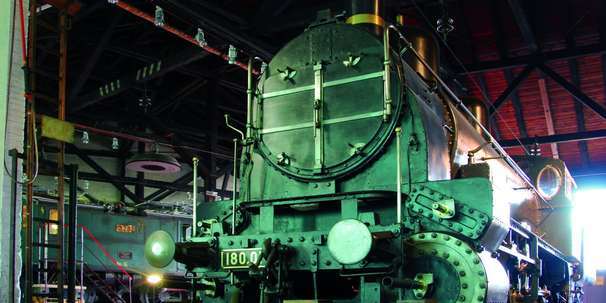 Eine große historische Dampflokomotive mit der Nummer 1800 ist in einem geräumigen Zugdepot im Industriestil mit einem hohen Dach und sichtbaren Balken ausgestellt.