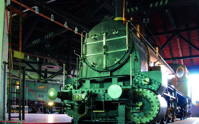 Eine große historische Dampflokomotive mit der Nummer 1800 ist in einem geräumigen Zugdepot im Industriestil mit einem hohen Dach und sichtbaren Balken ausgestellt.