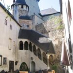 Mittelalterlicher Steinhof mit gewölbter Treppe, gelb karierten Tischen im Freien und einem burgähnlichen Gebäude im Hintergrund.