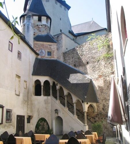 Mittelalterlicher Steinhof mit gewölbter Treppe, gelb karierten Tischen im Freien und einem burgähnlichen Gebäude im Hintergrund.