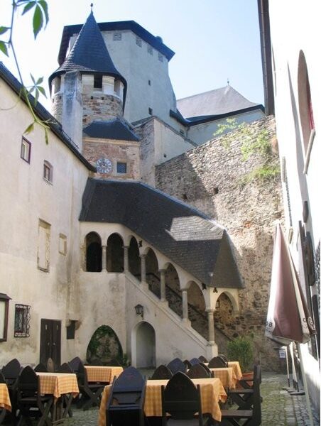 Mittelalterlicher Steinhof mit gewölbter Treppe, gelb karierten Tischen im Freien und einem burgähnlichen Gebäude im Hintergrund.