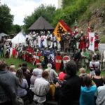 Eine Gruppe von Menschen in mittelalterlichen Kostümen mit Schilden und Fahnen steht im Freien, während eine Menschenmenge vor ihnen sitzt und zusieht.